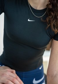 Femme portant un t-shirt noir Nike et un legging bleu Nike, avec un collier chaîne argenté et des cheveux bouclés sur l'épaule.
