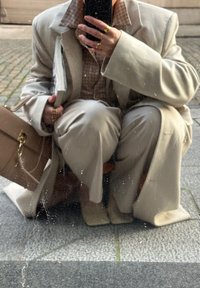 Beige oversized blazer en bijpassende broek van gladde stof. Een gestructureerd geruit shirt eronder, met een tijdschrift en een lichte handtas.