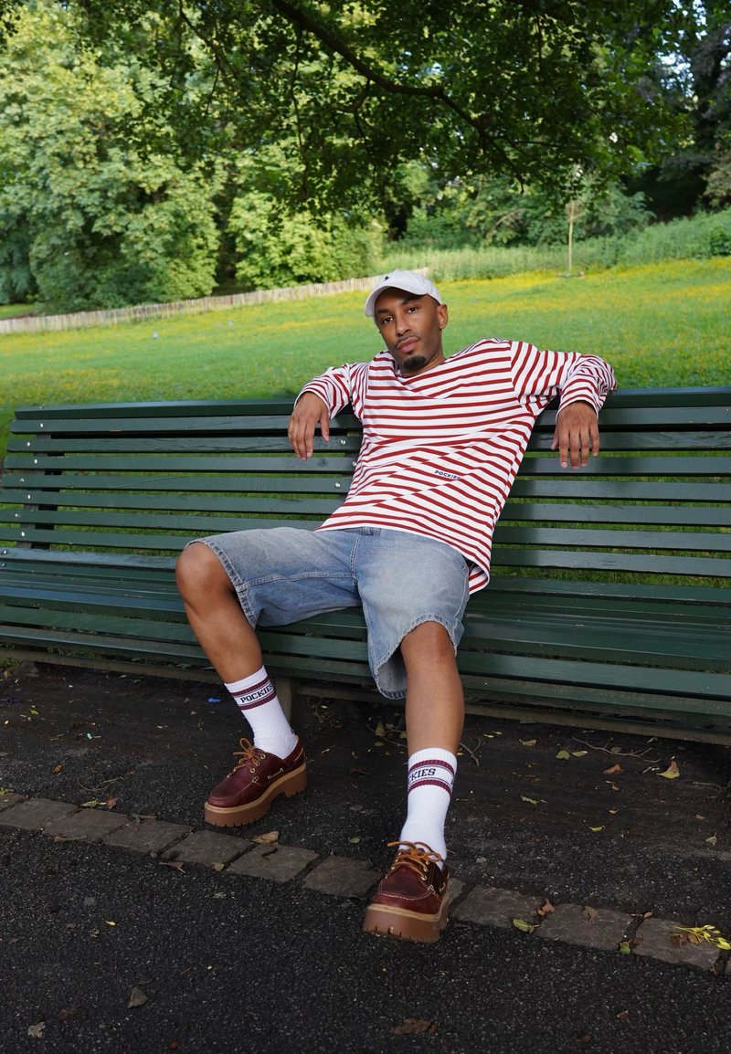 Homme portant une chemise rayée, un short en jean et une casquette blanche, assis détendu sur un banc vert dans un parc avec des arbres et de l’herbe en arrière-plan.