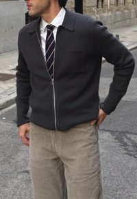 Veste zippée gris foncé avec col et poche poitrine, portée sur une chemise blanche et une cravate rayée, associée à un pantalon en velours côtelé beige clair.