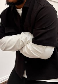 Chemise noire à boutons avec manches courtes portée par-dessus une chemise blanche à manches longues. Texture lisse, design minimaliste, sans motifs visibles.