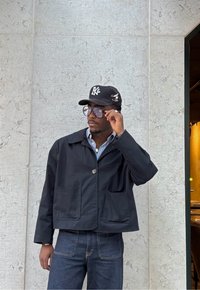 Crna kratka jakna s dva prednja džepa, nošena preko svijetloplave prugaste košulje, u kombinaciji s tamnim trapericama i crnom kapom.