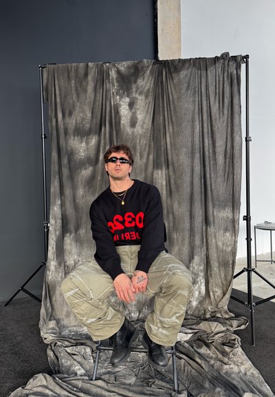 Suéter negro con texto rojo, pantalones cargo verdes y botas negras. El fondo cuenta con cortinas grises texturizadas y una pared sencilla.