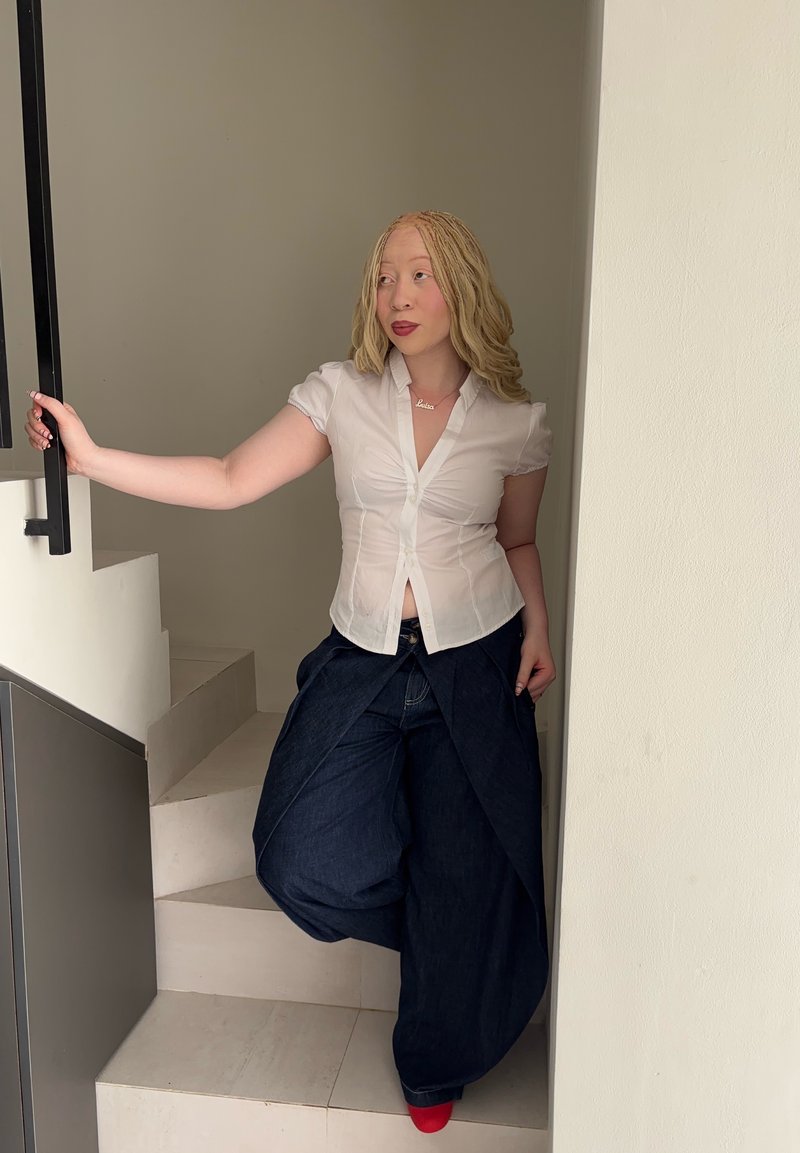 Young woman with braided blonde hair wearing white blouse, wide dark pants, and red shoes, leaning on stair railing in a neutral-toned interior.