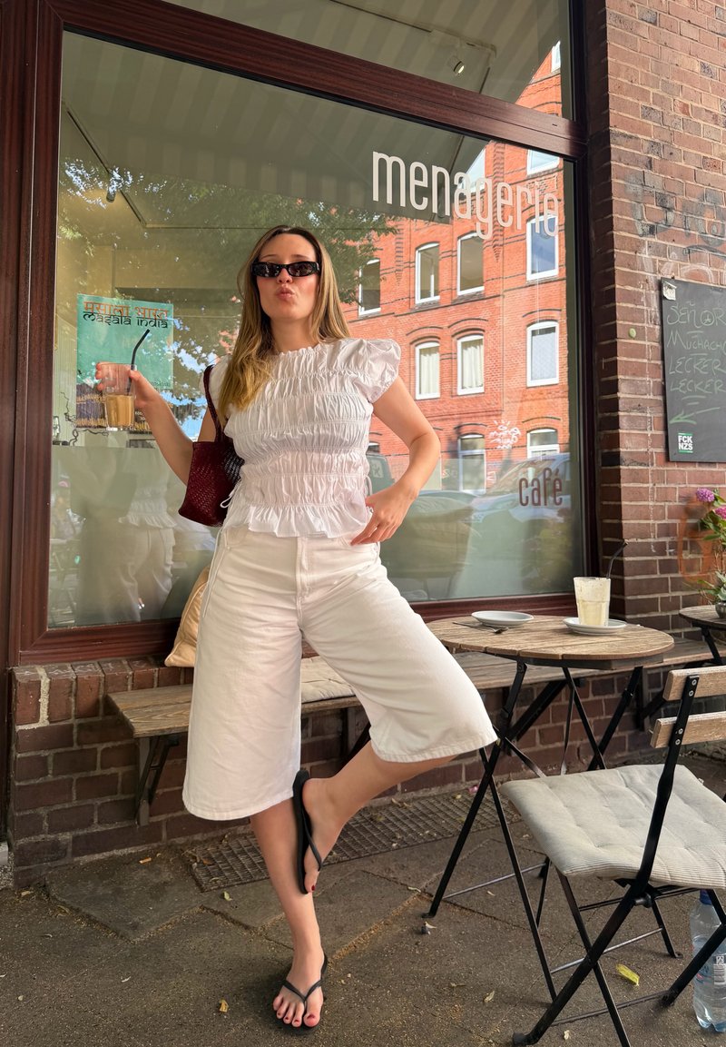 Femme en haut blanc et short, portant des lunettes de soleil, tenant une boisson glacée, debout avec une jambe levée devant un café avec des murs en briques et des tables extérieures.