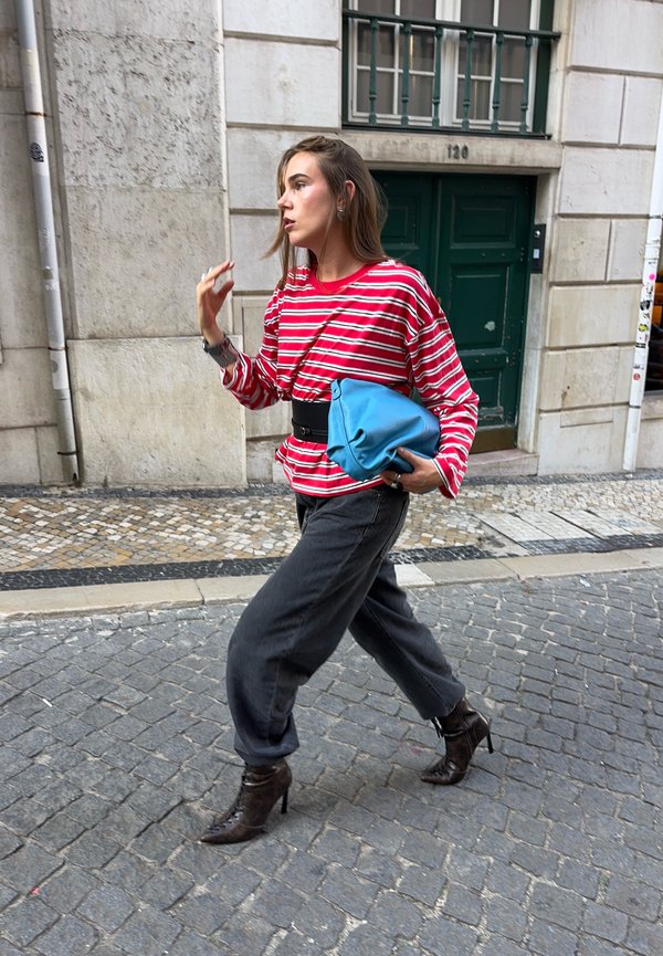 Mujer caminando por una calle de piedra, lleva una blusa roja a rayas, pantalones negros, botas de tacón alto y sostiene una bolsa azul doblada bajo un brazo.