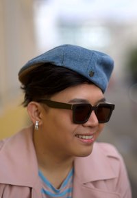 Beret din denim cu o formă rotunjită, ochelari de soare cu rame pătrate de culoare închisă și o jachetă roz deschis peste o bluză cu dungi. Un look texturat și casual.