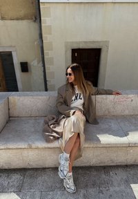 Blazer oversized bege, sweatshirt branco e uma saia de seda creme. Ténis brancos com detalhes cinzentos. Sentada em um banco de pedra texturizado.