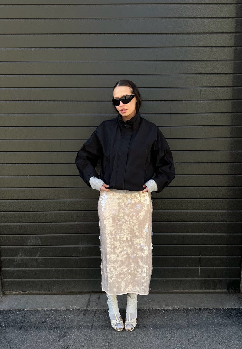 Femme portant une veste noire et des lunettes de soleil foncées, une jupe beige à sequins, des guêtres grises et des talons ouverts argentés, debout contre un mur sombre à panneaux.