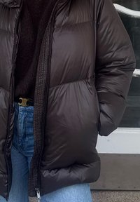 Doudoune noire avec une texture lisse et brillante, dotée d'un col montant, d'une fermeture éclair frontale et de sections matelassées horizontales. Portée par-dessus un pull sombre.