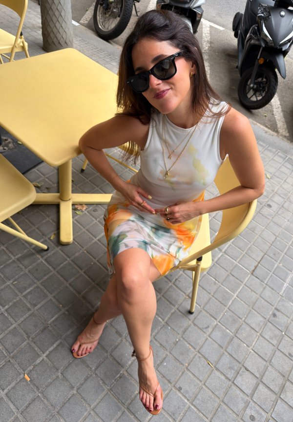 Žena so slnečnými okuliarmi a farebnými šatami sedí s prekríženými nohami na žltej stoličke v kaviarni vonku s kolobežkami v blízkosti.