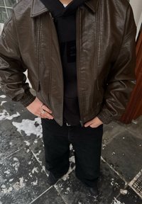 Veste aviateur en cuir marron avec fermeture éclair à l'avant, poignets côtelés et coupe courte, portée sur un sweat à capuche noir. Pantalon et chaussures noirs.