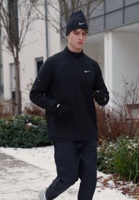 Jeune homme courant en plein air en hiver, portant une tenue de sport noire, des gants et un bonnet, avec de la neige et des plantes en arrière-plan.