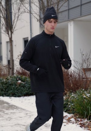 Joven corriendo al aire libre en invierno, vistiendo ropa deportiva negra, guantes y gorro, con nieve y plantas de fondo.