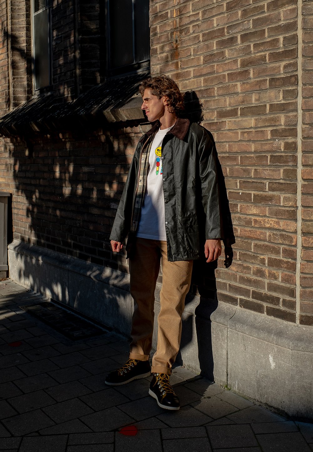 Mørkegrønn vanntett jakke med brun krage, hvit t-skjorte med grafikk, beige bukser og svarte støvler med gule snører. Solfylt urbant miljø.