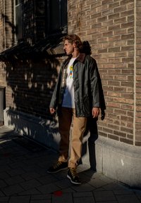 Mørkegrønn vanntett jakke med brun krage, hvit t-skjorte med grafikk, beige bukser og svarte støvler med gule snører. Solfylt urbant miljø.