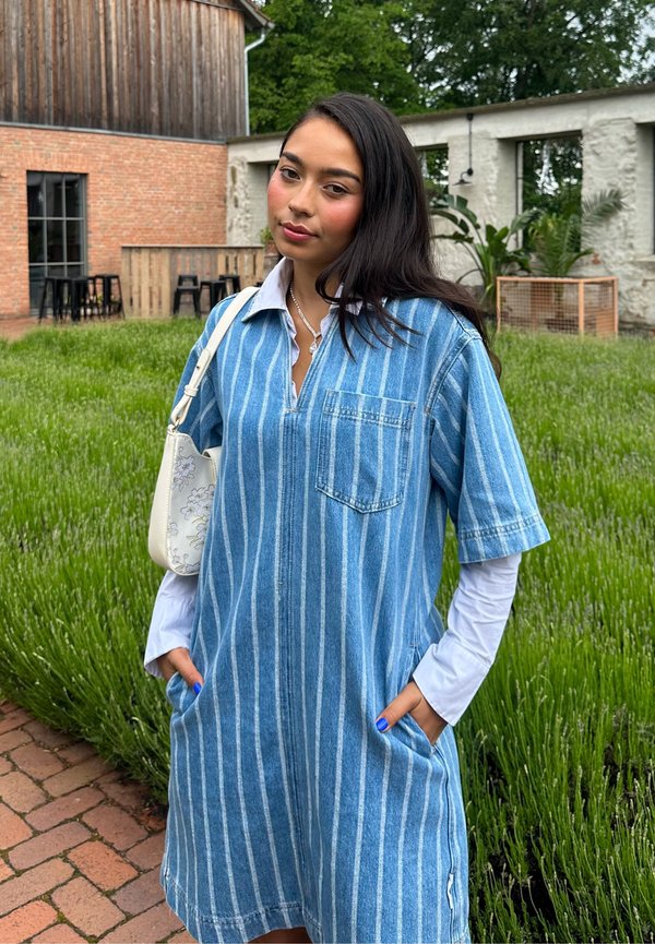 Junge Frau mit langen dunklen Haaren, die ein blau gestreiftes Kleid über einem weißen Langarmshirt trägt, steht in einem Garten mit grünen Pflanzen und Ziegelwegen.