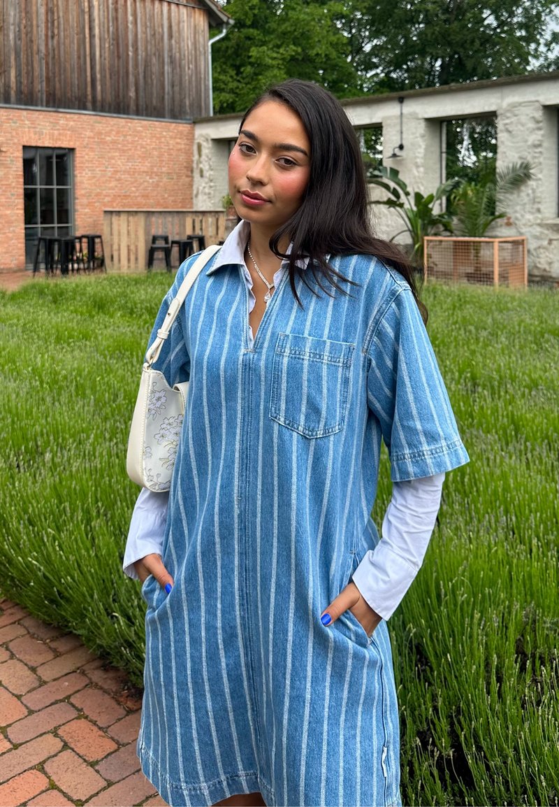 Jeune femme aux longs cheveux foncés portant une robe bleue rayée par-dessus une chemise blanche à manches longues, se tient dans un jardin avec des plantes vertes et des chemins en briques.