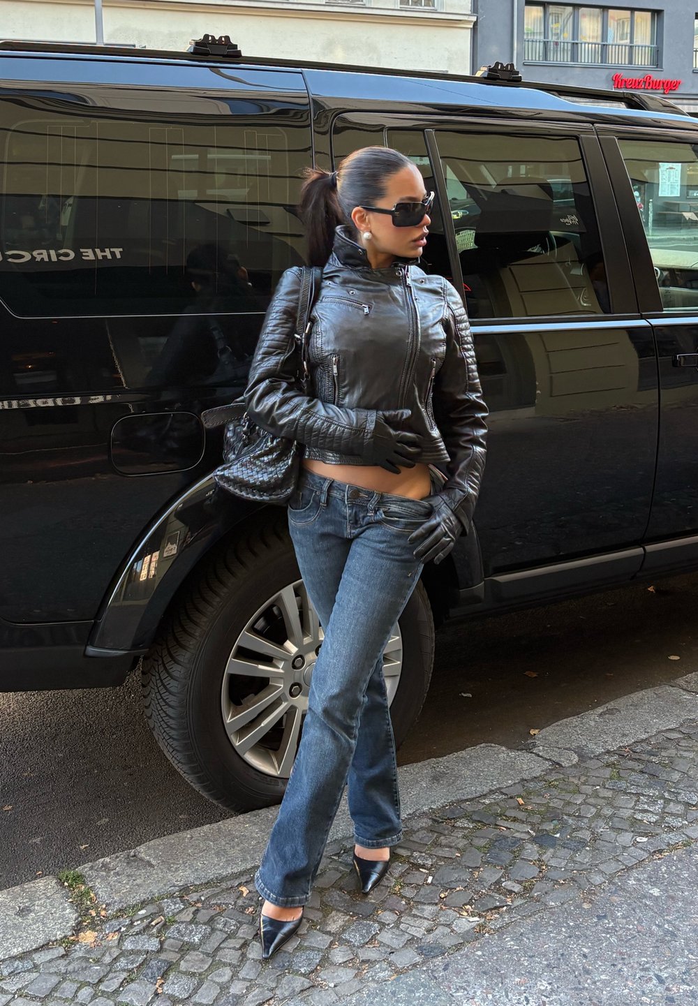 Schwarze Lederjacke, blaue Jeans, schwarze Handschuhe, hochhackige Schuhe, große schwarze Handtasche. Steht neben einem schwarzen Fahrzeug auf einer gepflasterten Straße.