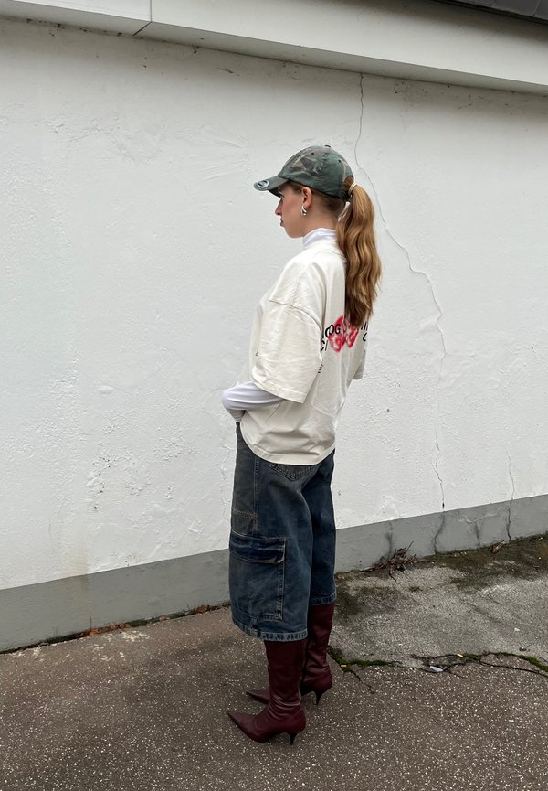 Oversized T-Shirt in crema con grafica rossa, indossato sopra un dolcevita bianco. Pantaloni cargo in denim blu e stivali a punta bordeaux.