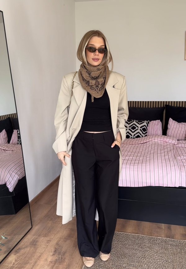 Mujer con un abrigo largo beige, pantalones negros, camiseta negra, bufanda marrón con estampado, gafas de sol y zapatos planos beige, de pie en un dormitorio.