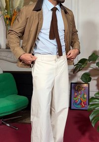 Veste marron clair avec col foncé, chemise bleu clair, cravate en tricot marron, et pantalon blanc à jambes larges. Chaise verte en arrière-plan avec une œuvre d'art.
