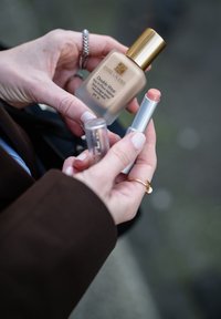 Estée Lauder Double Wear foundation, glassflaske med gullhett, og en sølv tube med en ferskenfarget applikator holdt i hånden.