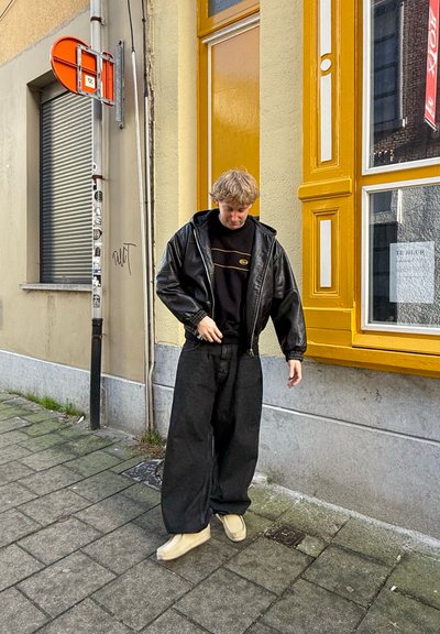 Persona con una chaqueta de cuero negra sobre un sudadera negra con acentos amarillos, vistiendo pantalones negros holgados y zapatos beige, de pie en una acera.