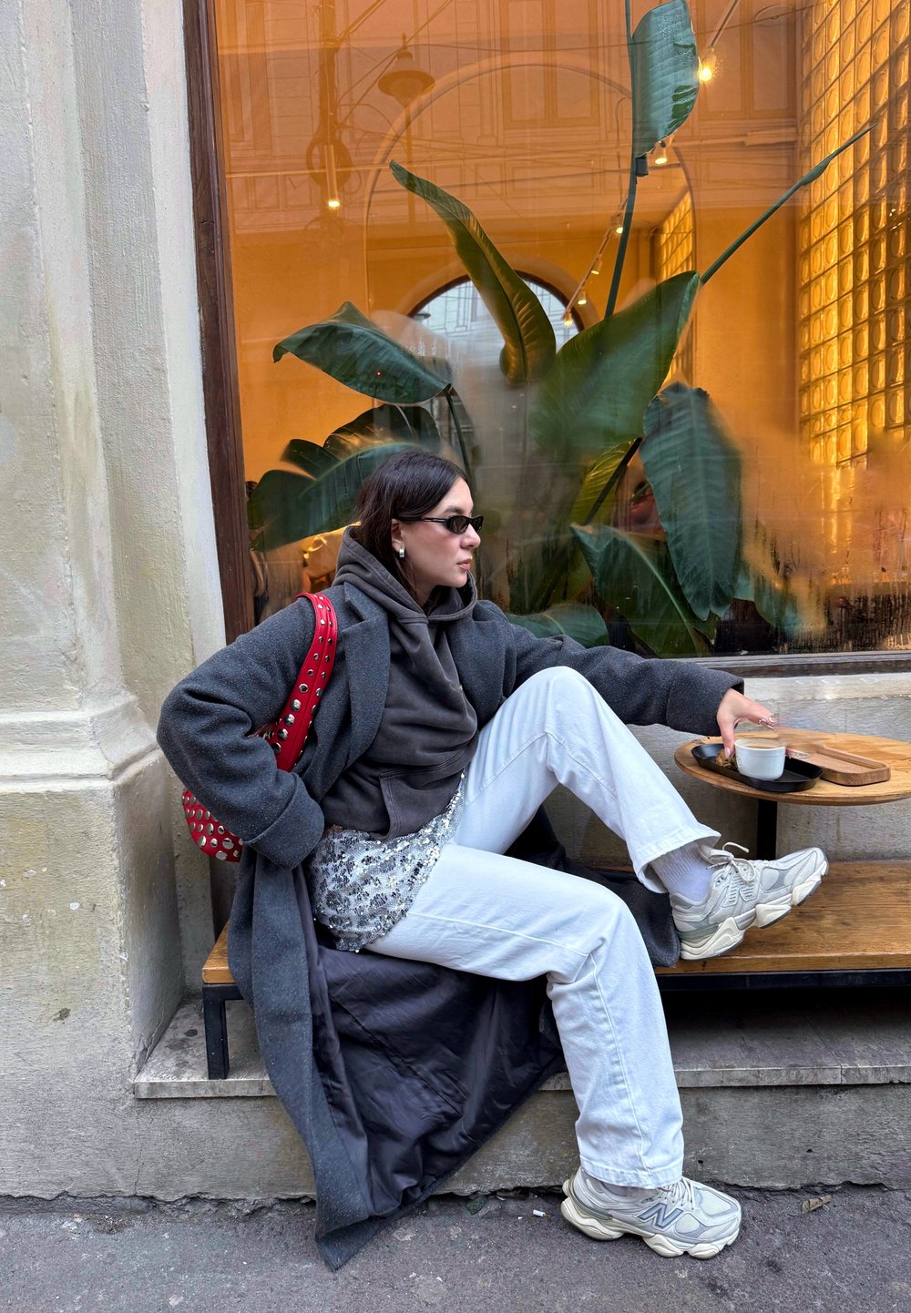 Une femme en long manteau gris est assise sur une plateforme en bois, portant un pantalon blanc et un sweat à capuche foncé, avec un sac rouge clouté et des baskets.