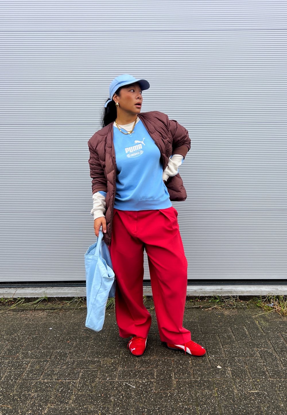 Casaco matelassê castanho sobre uma sweatshirt azul-claro com um logótipo, calças largas vermelhas, sapatos vermelhos e uma mala de mão azul-claro. Fundo neutro.