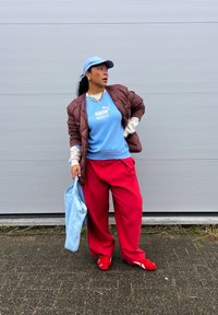 Casaco matelassê castanho sobre uma sweatshirt azul-claro com um logótipo, calças largas vermelhas, sapatos vermelhos e uma mala de mão azul-claro. Fundo neutro.