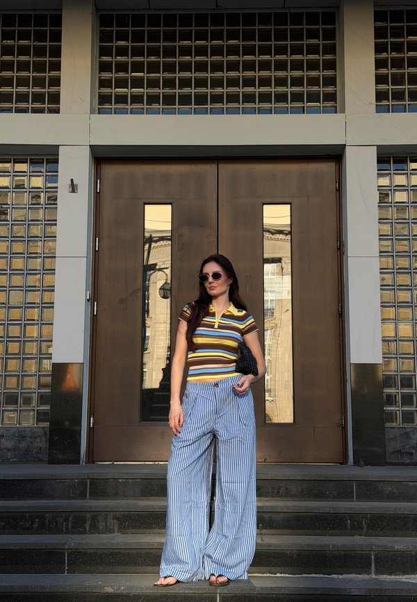 Femme portant des lunettes de soleil, une chemise rayée et un pantalon large rayé, debout sur des marches devant une grande porte double marron avec des panneaux en verre.