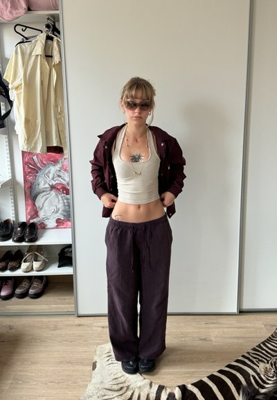 Chaqueta marrón sobre una camiseta sin mangas beige de canalé, combinada con pantalones sueltos morados oscuros. Los accesorios incluyen gafas de sol y collares en capas.