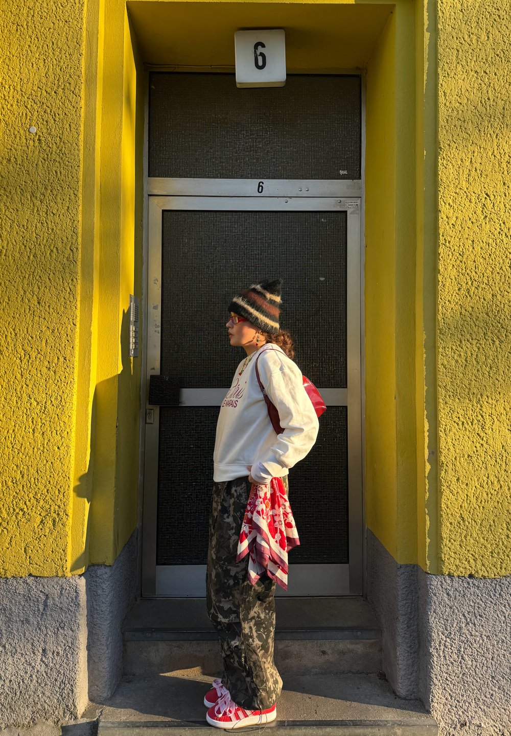 Sweatshirt blanc, pantalon camouflage, baskets rouges avec lacets, sac à dos rouge et écharpe à motifs. Debout à côté d'une porte verte avec un mur jaune.