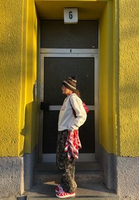 Sweatshirt blanc, pantalon camouflage, baskets rouges avec lacets, sac à dos rouge et écharpe à motifs. Debout à côté d'une porte verte avec un mur jaune.