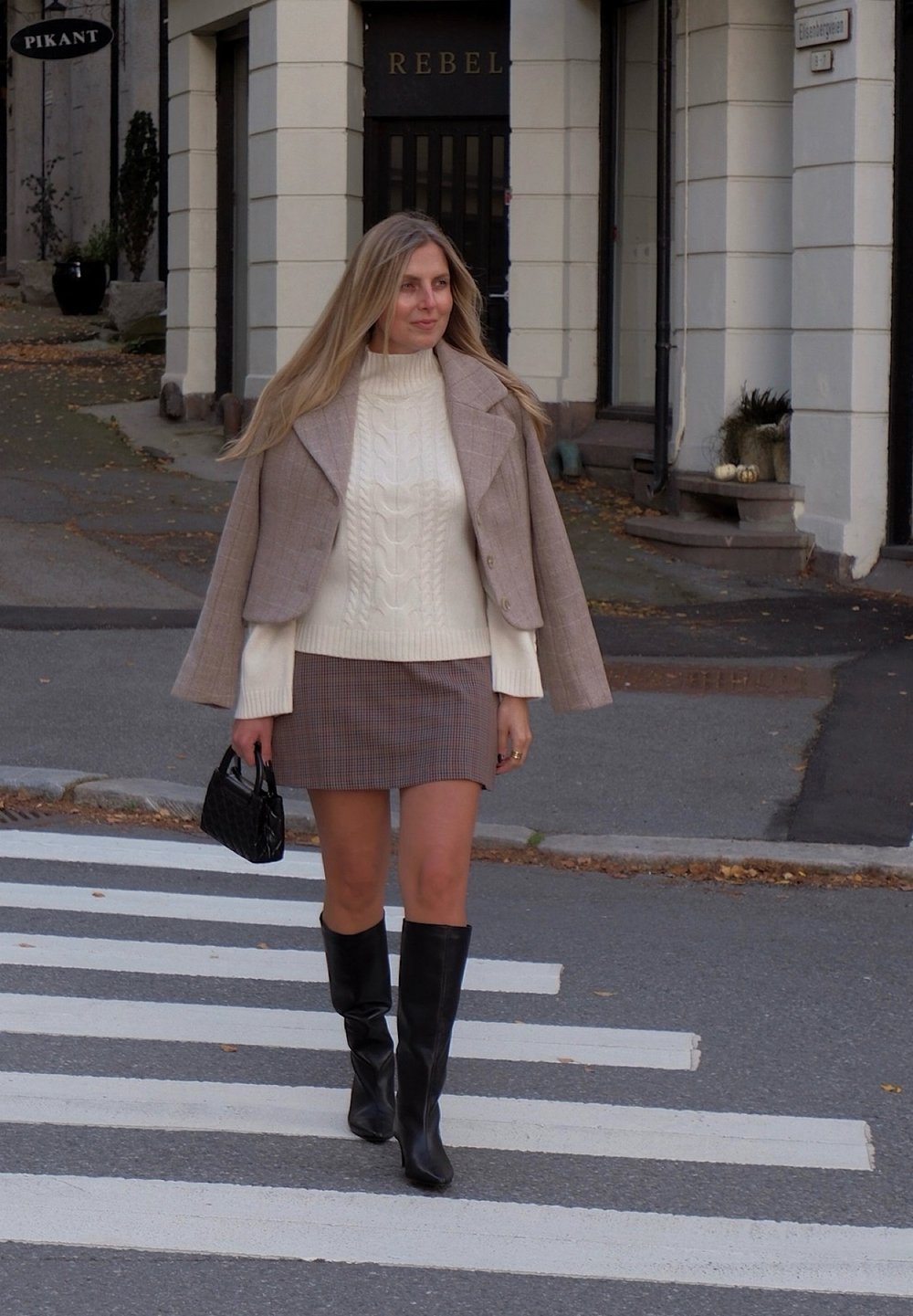 Beige geruite cropped jas over een witte kabelgebreide coltrui, gecombineerd met een geruite minirok en zwarte kniehoge laarzen. Zwarte handtas.
