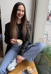 Young woman with long brown hair, wearing a black jacket and blue trousers, sitting by a window holding a coffee cup and smiling.