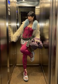 Personne en tenue rose à lacets, sandales plateformes blanches et veste duveteuse se tient dans un ascenseur métallique en tenant un sac violet.