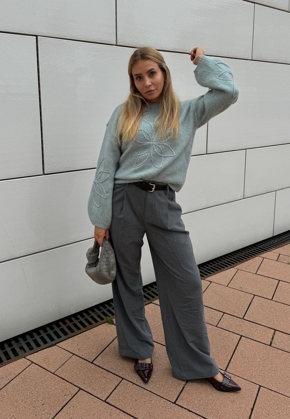 Vaaleansininen neulottu neulepusero, jossa on kukkakirjontaa, yhdistetty harmaisiin leveälahkeisiin housuihin ja viininpunaisiin kärjenmuotoisiin korkokenkiin. Kudottu harmaa käsilaukku.