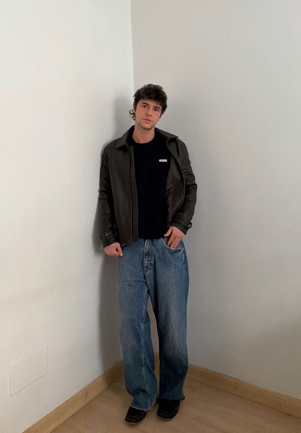 Jeune homme aux cheveux foncés debout dans un coin, portant une veste noire, une chemise noire, un jean bleu ample et des chaussures noires sur un sol en bois.