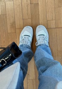 Baskets blanches avec des accents gris, présentant une empeigne texturée et des lacets. Portées avec un jean bleu clair. Sac à main noir partiellement visible.