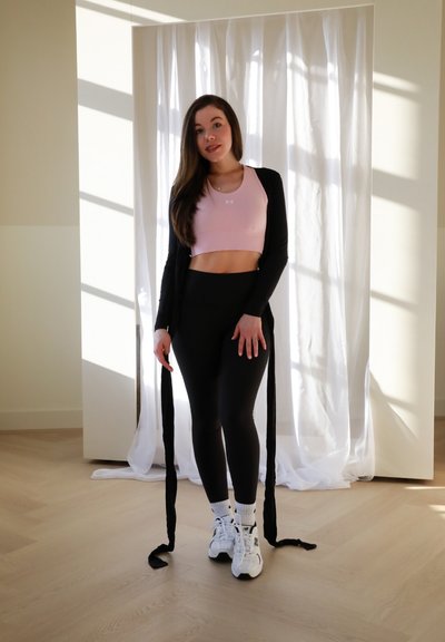 Conjunto atlético con un top corto rosa, leggings negros y un cárdigan largo negro. Zapatillas blancas y suelo de madera texturizada visible.