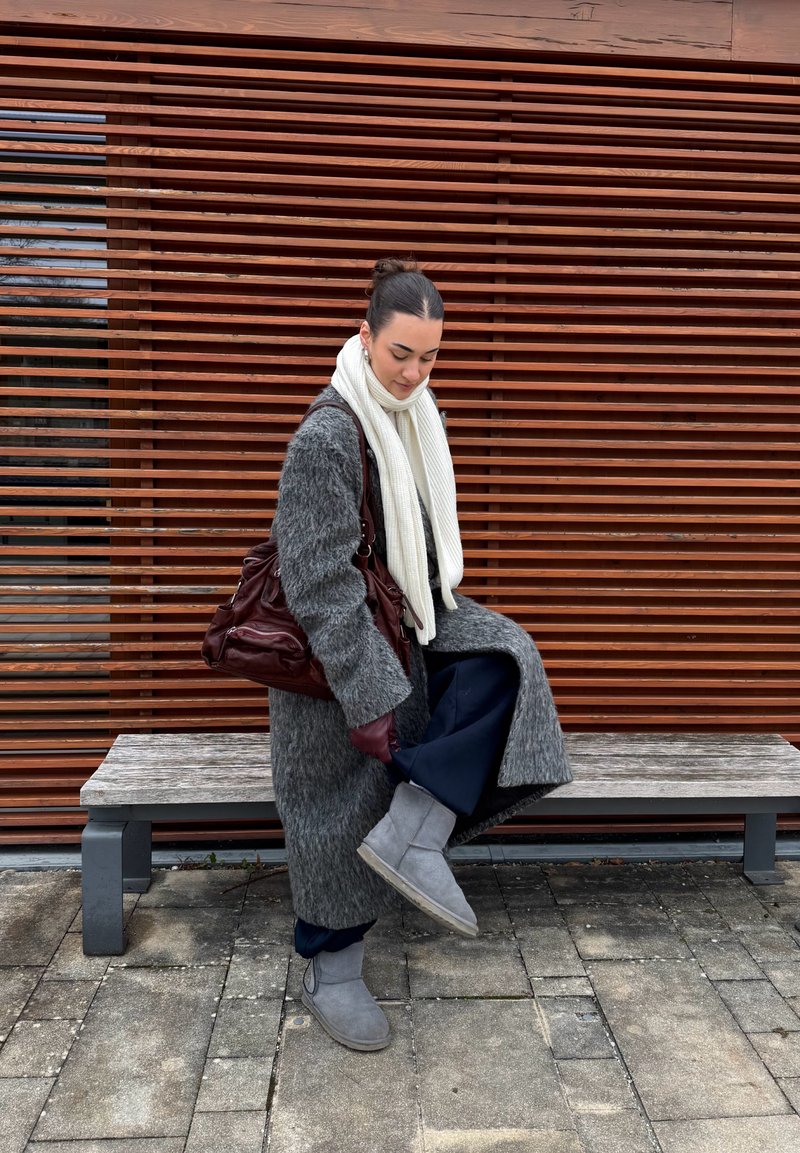 Szary, puszysty płaszcz z szerokimi rękawami, biały dziergany szalik, granatowe spodnie i szare krótkie botki. Torebka w ciemnoczerwonym kolorze. Drewniane tło z listew.