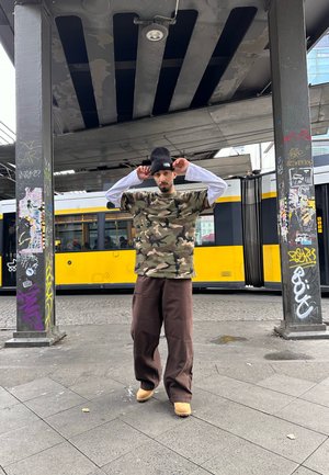 Kamufliažo marškinėliai trumpomis rankovėmis, derinami su laisvais rudi kelnėmis. Asmuo dėvi juodą skrybėlę ir stovi po urbanistine struktūra.
