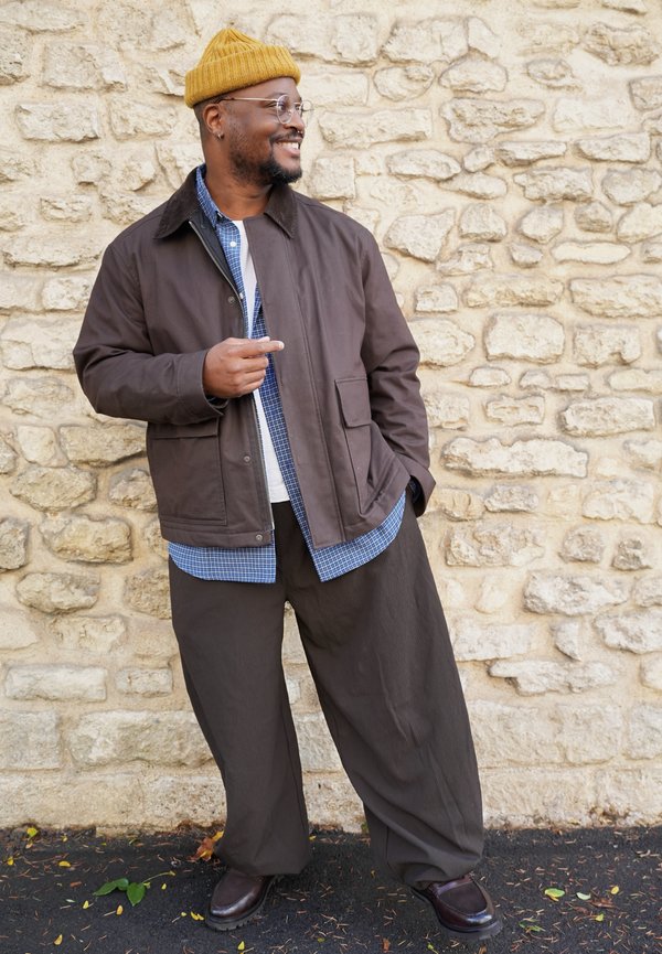 Un homme souriant, coiffé d’un bonnet moutarde et portant des lunettes, une veste brune et un pantalon ample, se tient près d’un mur de pierre et montre le côté.