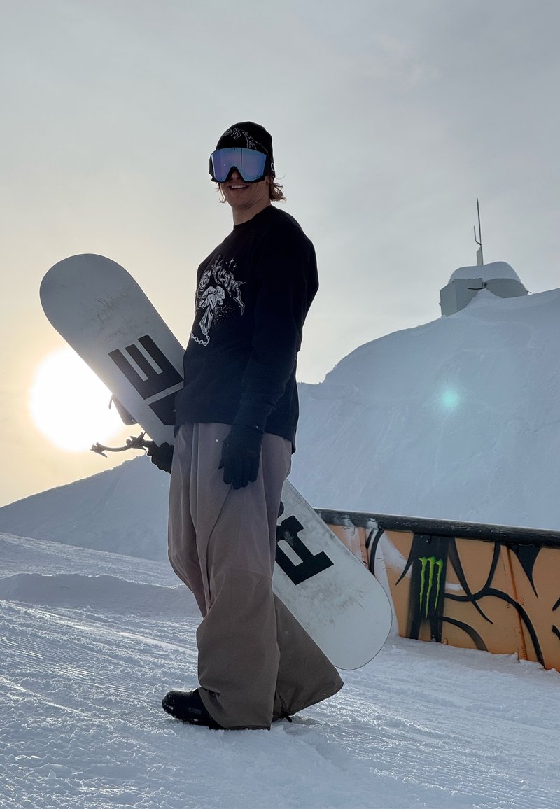 Persona sniega apģērbā ar snovbordu rokās, stāv uz sniegotas nogāzes pie margas ar Monster logo, saulē zemu debesīs aiz viņiem.