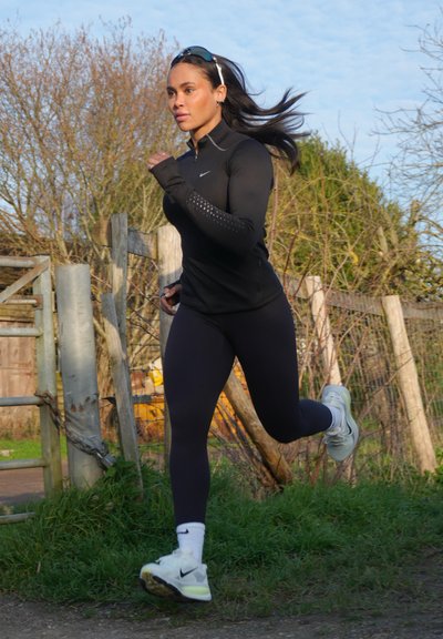 Mujer con cabello largo y oscuro corriendo al aire libre por un camino de tierra, vestida con ropa deportiva negra, zapatillas blancas y gafas de sol en la cabeza.