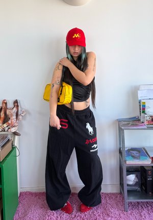 Mujer con cabello largo que lleva una gorra roja, top negro sin mangas, pantalones negros anchos con logotipos y zapatos rojos, sosteniendo un bolso amarillo de hombro en interiores.