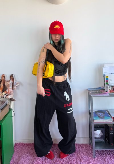 Mujer con cabello largo que lleva una gorra roja, top negro sin mangas, pantalones negros anchos con logotipos y zapatos rojos, sosteniendo un bolso amarillo de hombro en interiores.