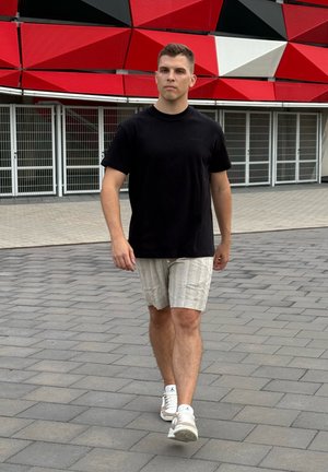 Hombre caminando sobre un pavimento de baldosas, vistiendo una camiseta negra de manga corta y pantalones cortos de rayas claras, combinados con zapatillas blancas.